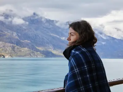 Eine Frau im blau-karierten Mantel blickt an Bord der Nieuw Amsterdam auf die neblige Berglandschaft von Alaska.
