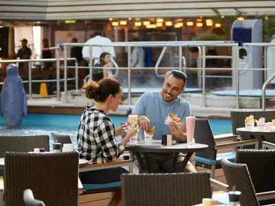 Ein lächelndes Paar sitzt an Bord der Nieuw Amsterdam an einem Tisch am Pool und isst Burger und Pommes vom Dive In.