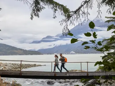Ein Paar wandert in Alaska über eine Brücke vor der Kulisse von Bergen, Wolken und der im Hintergrund liegenden Nieuw Amsterdam.
