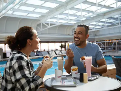 Ein lachendes Paar beim Mittagessen mit Burger, Pommes und Milchshake am Pool an Bord der Nieuw Amsterdam.