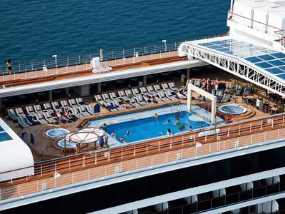 Vogelperspektive auf den sonnigen Lido-Poolbereich an Bord der Nieuw Amsterdam mit Liegestühlen und Whirlpools direkt am Meer.