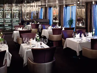 Ein luxuriös eingerichtetes Restaurant an Bord der Nieuw Amsterdam mit weiß eingedeckten Tischen und bequemen Ledersesseln.