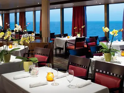 Eingedeckte Tische in einem eleganten Restaurant an Bord der Nieuw Amsterdam mit großen Fenstern und Blick auf das blaue Meer.