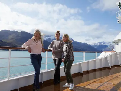 Drei lächelnde Frauen auf dem Außendeck an Bord der Nieuw Amsterdam vor der Kulisse schneebedeckter Berge in Alaska.