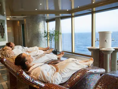 Vier Frauen entspannen in weißen Bademänteln auf beheizten Mosaik-Liegen im Greenhouse Spa an Bord der Nieuw Amsterdam mit Blick auf das Meer.