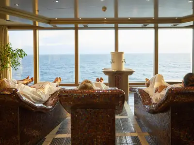 Gäste entspannen auf beheizten Mosaik-Liegen im Spa-Bereich an Bord der Nieuw Amsterdam und blicken durch Panoramafenster auf das weite Meer.