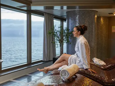 Eine Frau im Bademantel entspannt auf einer beheizten Liege im Spa-Bereich an Bord der Nieuw Amsterdam und blickt durch die Fenster auf das Meer.
