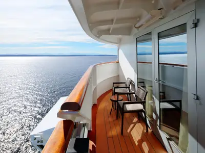 Die private Holzdeck-Veranda einer Suite an Bord der Nieuw Amsterdam bietet zwei Sessel, einen Tisch und einen weiten Blick über das sonnige Meer.