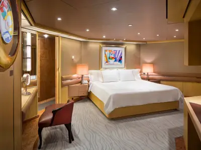 Die geräumige Pinnacle Suite an Bord der Nieuw Amsterdam mit großem Doppelbett, zwei Nachttischlampen, Kunstwerk und stilvollem Sessel.