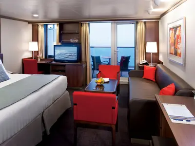 Blick in eine geräumige Suite an Bord der Nieuw Amsterdam mit Bett, Sitzecke mit roten Sesseln und Sofa sowie Balkon mit Meerblick.