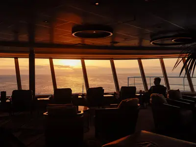 Blick durch die Panoramafenster der Crow's Nest Lounge an Bord der Nieuw Statendam auf einen goldenen Sonnenuntergang über dem Meer.
