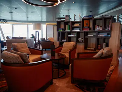 Bequeme Sessel und ein kleiner Tisch in der gemütlichen Crow's Nest Lounge an Bord der Nieuw Statendam, im Hintergrund ein Bücherregal.
