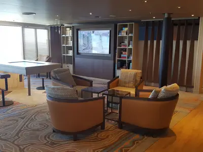 Einladender Loungebereich an Bord der Nieuw Statendam mit bequemen Sesseln, gemustertem Teppich, Bücherregalen und einem großen Monitor.