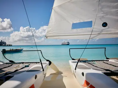 Blick von zwei am Strand liegenden Katamaranen über das türkisfarbene Meer auf die entfernte Nieuw Statendam.