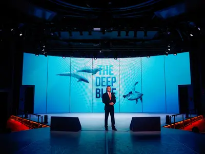 Ein Redner im dunklen Anzug steht auf der Bühne der World Stage an Bord der Nieuw Statendam vor einer LED-Wand.