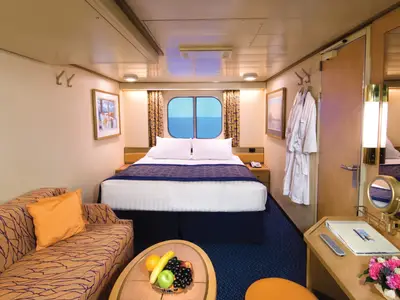 Blick in eine komfortable Außenkabine an Bord der Noordam mit Doppelbett, Sofa, Obstteller und einem Fenster mit Meerblick.