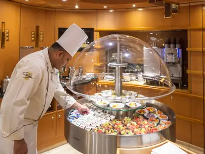 Ein Koch in weißer Uniform arrangiert Obst und Joghurt an einer runden Buffetstation in der edlen Neptune Lounge an Bord der Noordam.