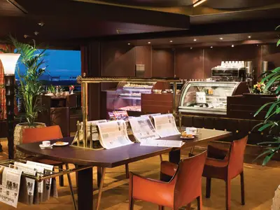Innenansicht des warm beleuchteten Explorations Café an Bord der Noordam mit Ledersesseln, Zeitungsständern und einer Kaffeebar.
