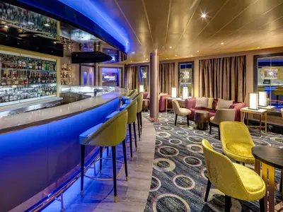 Elegant beleuchtete Bar-Lounge an Bord der Noordam mit blauem Tresenlicht, stilvollen gelben Sesseln und Barhockern vor einer Spirituosenwand.