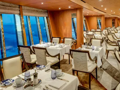 Elegant eingedeckte Tische und weiße Stühle im Hauptrestaurant an Bord der Noordam mit Blick auf das Meer bei Dämmerung.