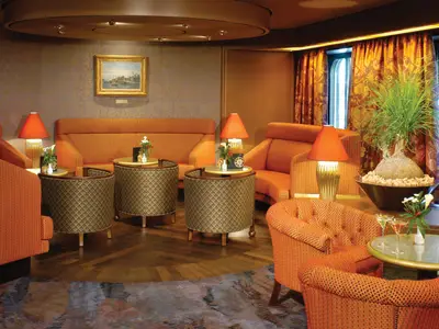 Die elegant eingerichtete Explorer's Lounge an Bord der Oosterdam mit gemütlichen orangefarbenen Polstermöbeln, Tischlampen und Fensterblick.