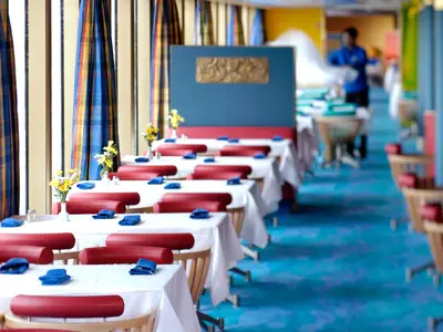 Hell erleuchtetes Restaurant an Bord der Oosterdam mit weiß gedeckten Tischen, roten Polstern und blauem Teppich.