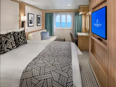 Blick in eine moderne Außenkabine an Bord der Rotterdam mit Doppelbett, Sofa, Fernseher und Meerblick durch das Fenster.