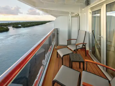 Balkon einer Kabine an Bord der Rotterdam mit zwei Stühlen, Fußbänken und Tisch sowie Ausblick auf einen bewachsenen Wasserweg.