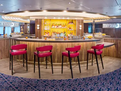 Elegante Half Moon Bar an Bord der Rotterdam mit einer geschwungenen Holztheke, roten Barhockern und gelb beleuchteten Regalen voller Flaschen.