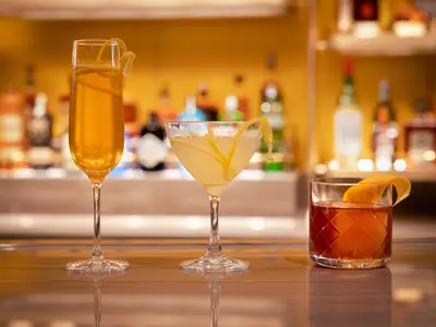Drei stilvoll angerichtete Cocktails mit Zitrusgarnitur stehen auf dem Tresen einer Bar an Bord der Rotterdam vor einem warm beleuchteten Hintergrund.