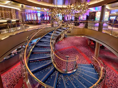 Blick auf die geschwungene Wendeltreppe mit blauem Teppich und das angrenzende Casino an Bord der Rotterdam.