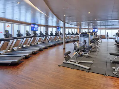Blick in das moderne Fitness Center an Bord der Rotterdam mit Laufbändern vor den Fenstern und Kraftgeräten auf dem Sportboden.