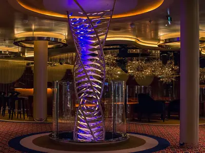 Blick auf eine spiralförmige, blau beleuchtete Kunstskulptur in einer eleganten Lounge an Bord der Rotterdam mit Barhockern im Hintergrund.