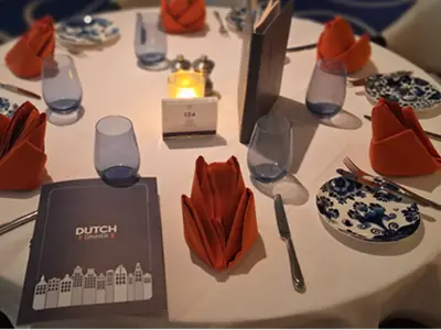 Festlich gedeckter runder Tisch an Bord der Rotterdam mit orangefarbenen Servietten, blau-weißem Geschirr und Menükarten für das Dutch Dinner.