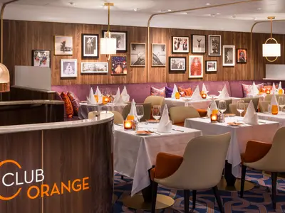 Elegantes Restaurant Club Orange an Bord der Rotterdam mit weiß eingedeckten Tischen, beigen Sesseln und einer Wand mit gerahmten Fotos.