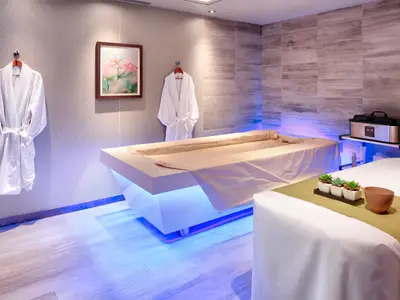 Ein Spa-Behandlungsraum an Bord der Rotterdam mit einer beleuchteten Liege, Massagetisch, Bademänteln und entspannter Atmosphäre.