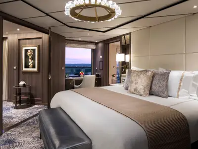 Blick auf das luxuriöse Doppelbett und den Schreibtisch in der Pinnacle Suite an Bord der Rotterdam mit Abendhimmel im Fenster.