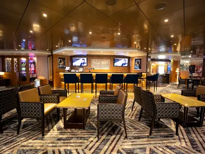 Eine stilvolle Lounge an Bord der Volendam mit bequemen Sesseln, gelben Tischen, Barhockern an der Theke und Bildschirmen.