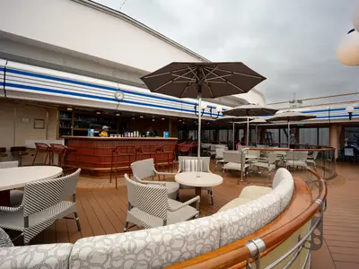 Die Lido Bar an Bord der Volendam mit gemütlichen Sesseln, Holzboden und Sonnenschirmen unter bewölktem Himmel.
