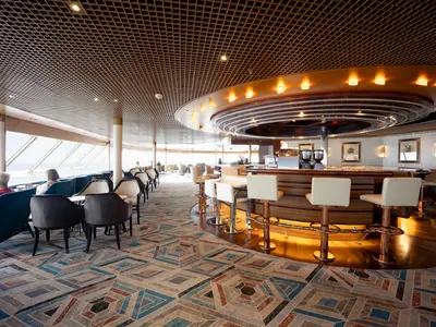 Die stilvolle Crow's Nest Lounge an Bord der Volendam mit einer kreisförmigen Bar, Barhockern und Panoramafenstern mit Blick auf das Meer.