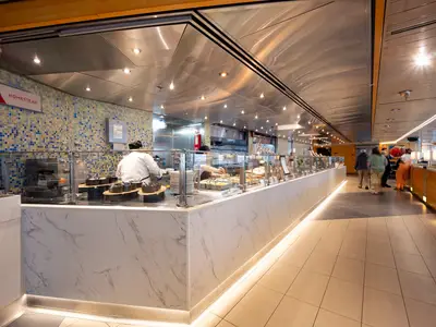 Das Buffet-Restaurant an Bord der Volendam mit einer langen Theke, Köchen hinter Glasscheiben und Gästen auf der rechten Seite.