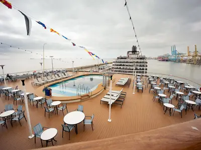 Der Sea View Poolbereich auf dem Achterdeck an Bord der Volendam mit Liegestühlen, Tischen und Blick auf das Meer und einen Hafen.