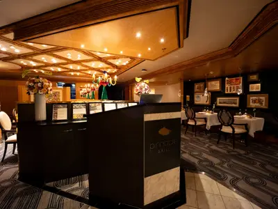 Der Empfangstresen des Pinnacle Grill Restaurants an Bord der Volendam mit eleganten Tischen, Gemälden und warmer Beleuchtung im Hintergrund.
