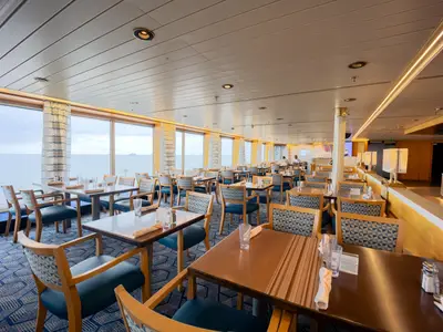 Das klassisch eingerichtete Restaurant an Bord der Volendam mit blauem Teppichboden und großen Panoramafenstern mit Blick aufs Meer.