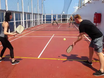 Vier Personen beim Pickleball-Spiel auf dem roten Außensportdeck an Bord der Volendam bei Sonnenschein.
