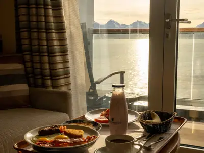 Frühstück in der Kabine an Bord der Westerdam mit Blick durch die Balkontür auf das glitzernde Meer und schneebedeckte Berge in Alaska.
