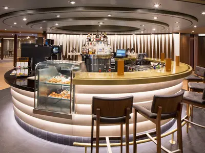 Blick auf die stilvoll beleuchtete, runde Bar im Explorations Café an Bord der Westerdam mit Gebäckvitrine und Barhockern.
