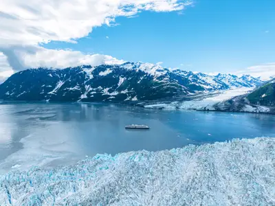 Vogelperspektive der Westerdam in einer Bucht in Alaska, umgeben von eisigen Gletschern und schneebedeckten Bergen unter blauem Himmel.