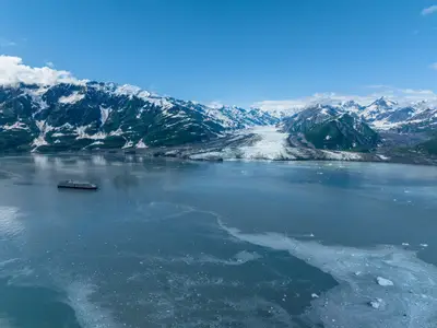 Luftaufnahme der Westerdam vor einer majestätischen Bergkette mit schneebedeckten Gipfeln und einem riesigen Gletscher in Alaska.