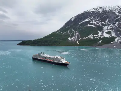 Luftaufnahme der Westerdam, die in türkisblauem Wasser voller Treibeis vor einer verschneiten Bergkette in Alaska fährt.